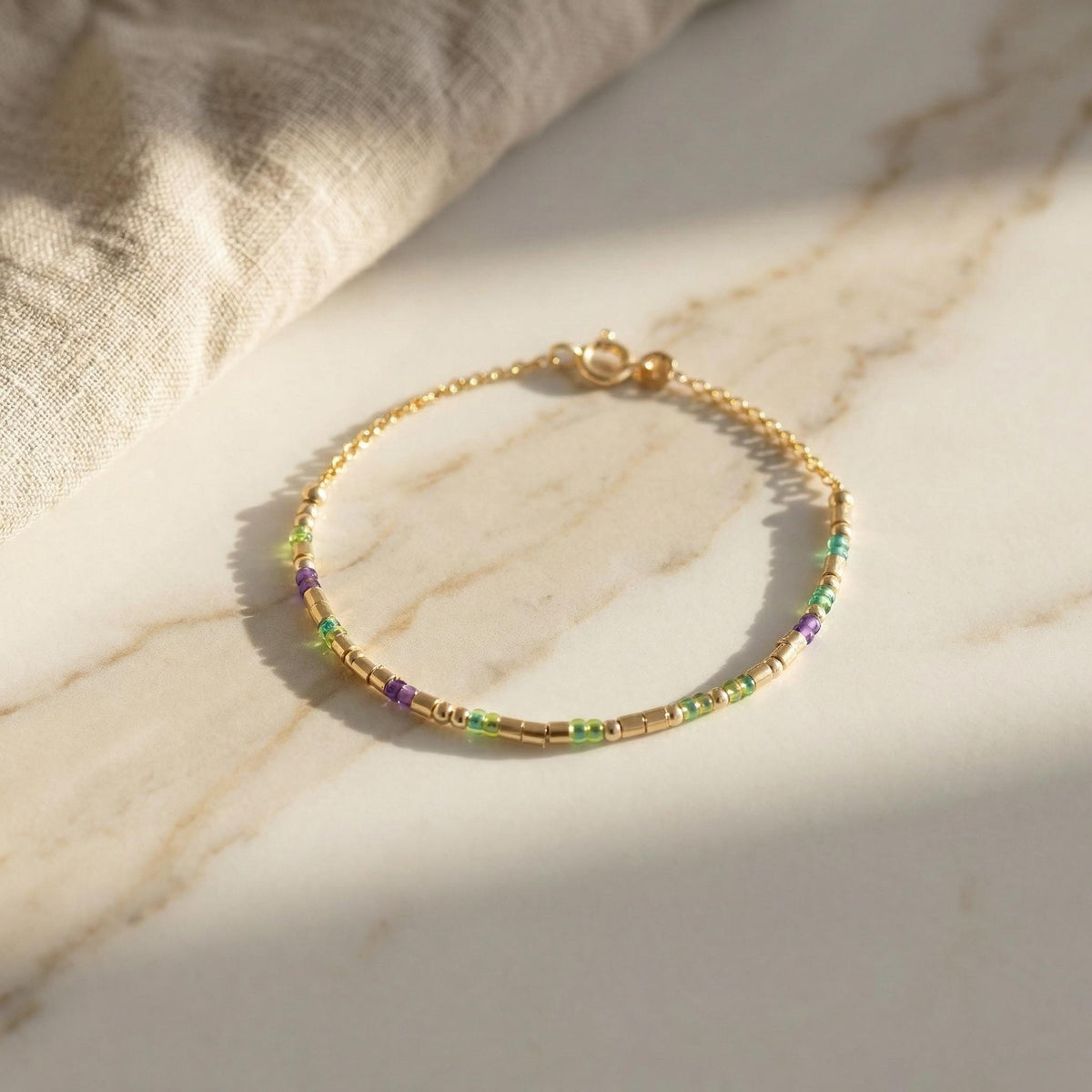 14k gold fill morse code bracelet with green and purple glass beads, angled on marble and linen
