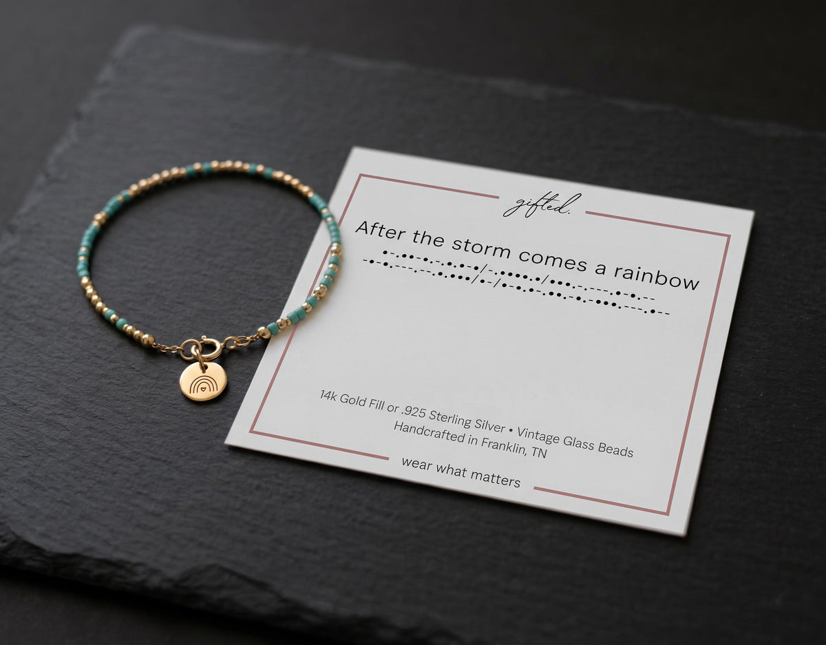 Morse code bracelet in 14k Gold Fill with the rainbow charm, resting on the printed card on dark slate